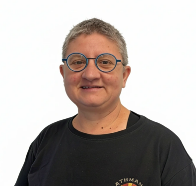 Portrait of a person with short gray hair and round blue glasses, wearing a black t-shirt with a Kathmandu logo and 'Since 1997' text.